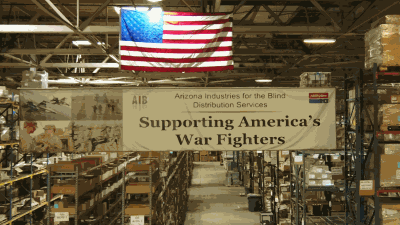 A picture of the AIB DSU Warehouse with racking in the background and a banner in the foreground the reads: Supporting America's War Fighters